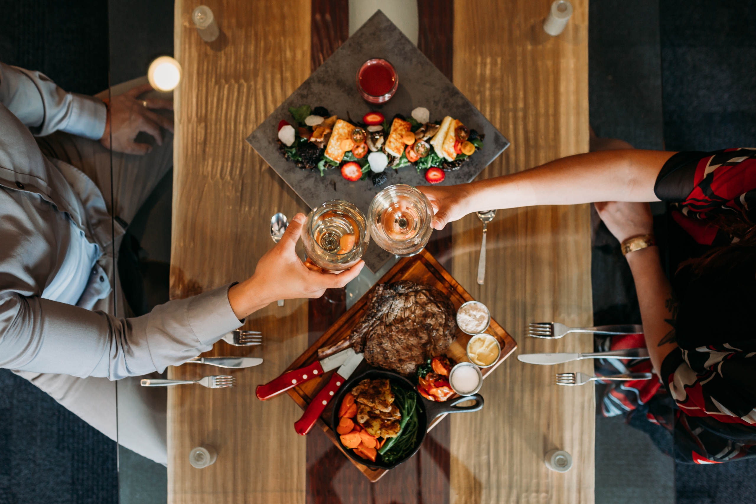 Aerial view of a table with plates of food and two people cheers-ing a glass of wine