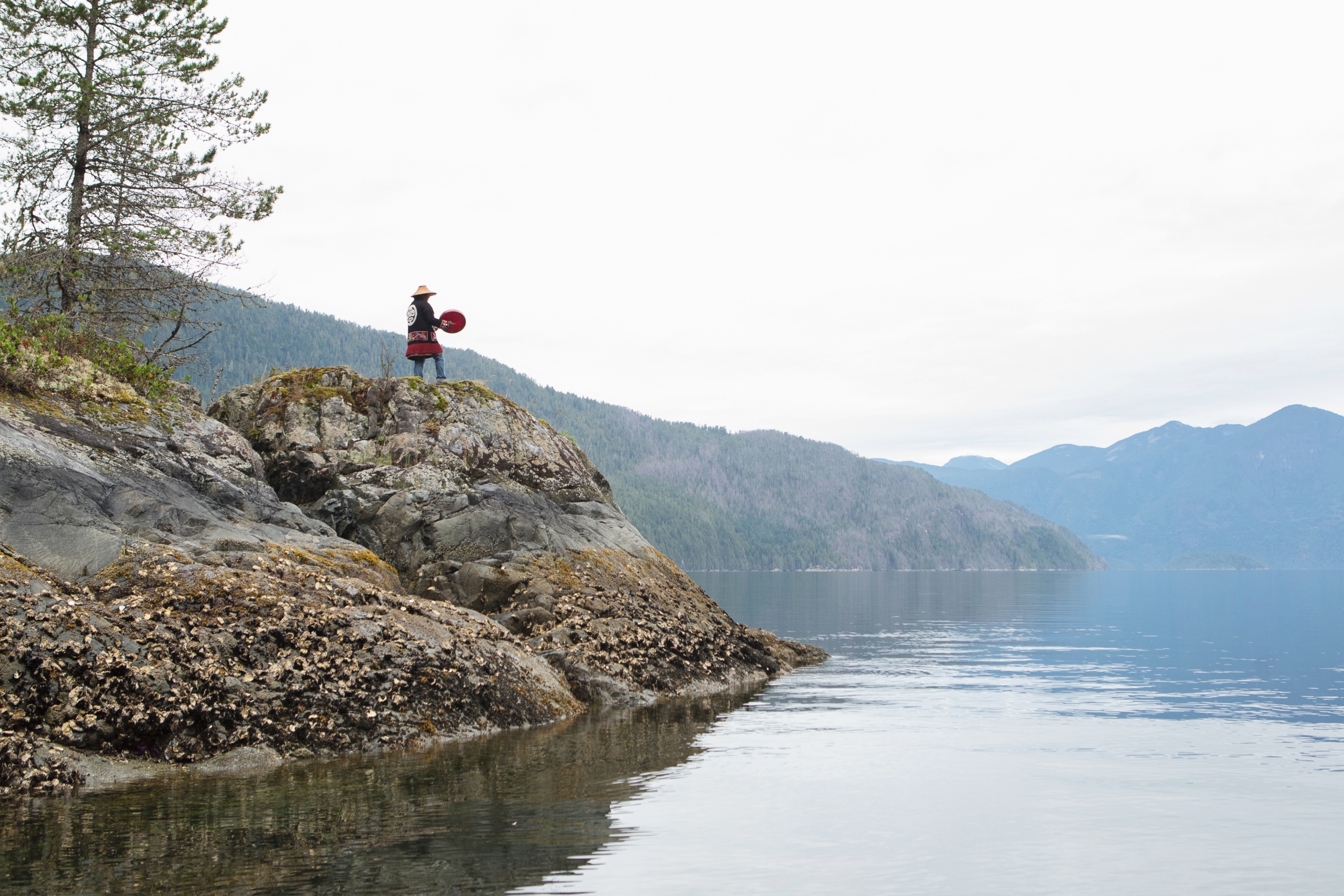 A person drumming standing on the edge of a rock overlooking the water