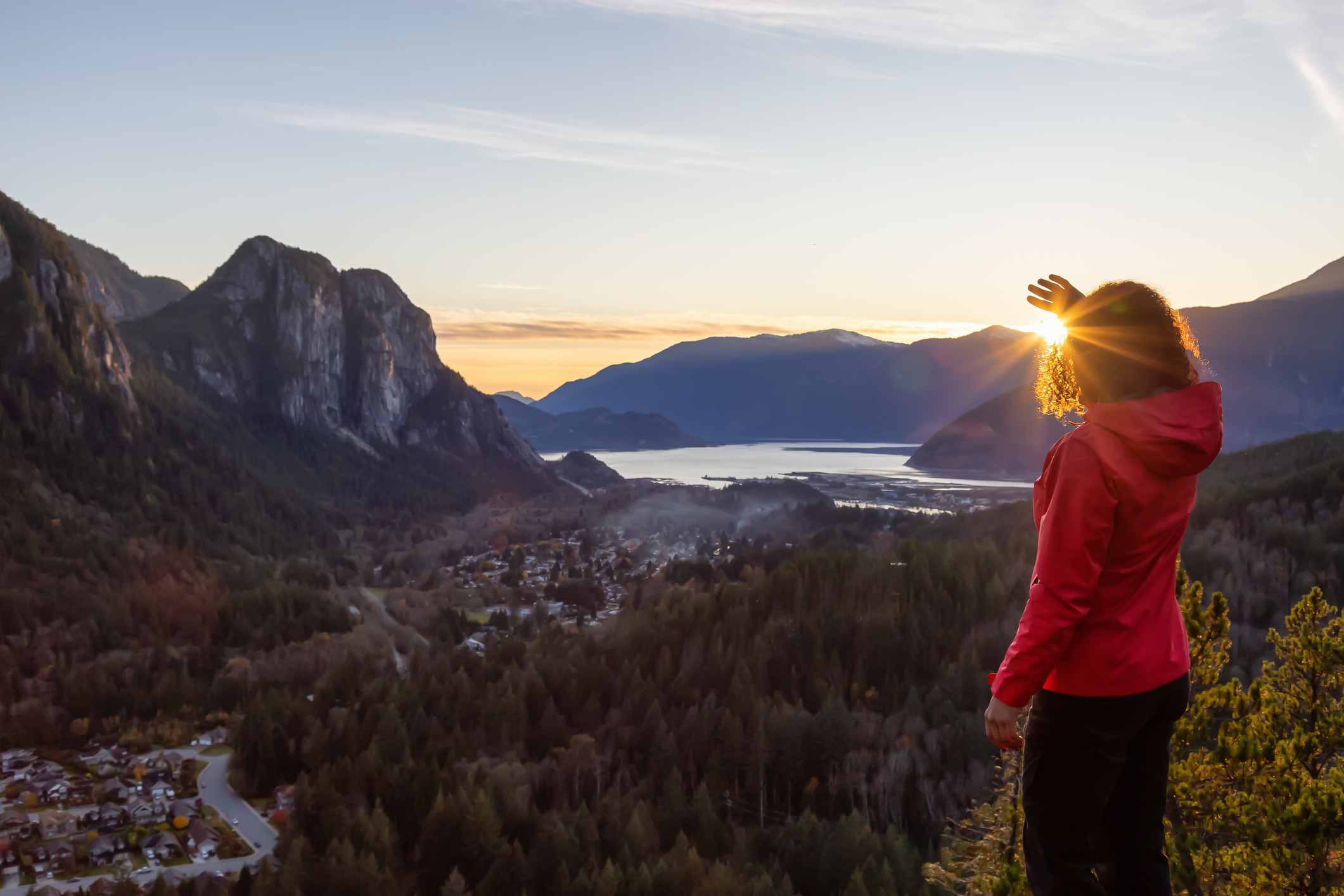 Woman standing on a cliff looking at water and mountains at sunset