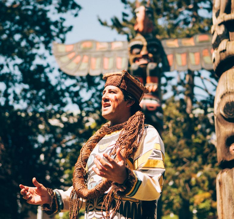 A man wearing traditional Indigenous clothing stands in front of two totem poles