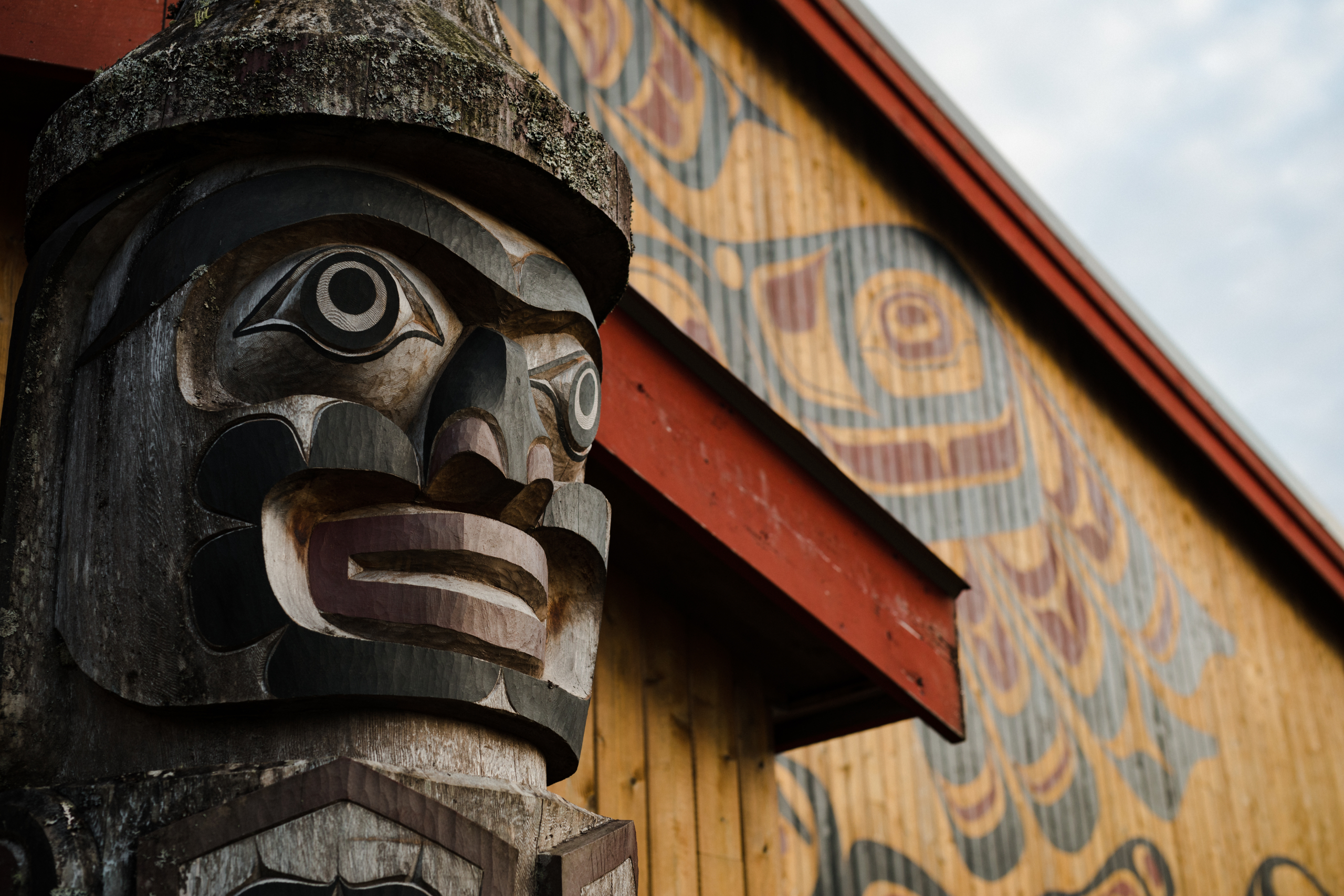 Up close image of a carved face on a totem pole and painted exterior of a building.