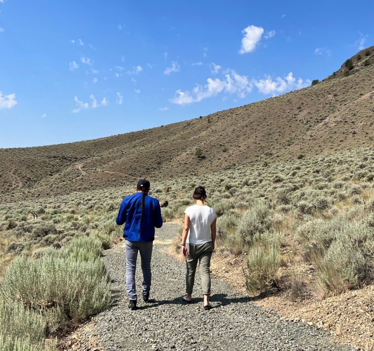 Two people hiking on a gravel trail through shrubby hills under a blue sky with clouds.