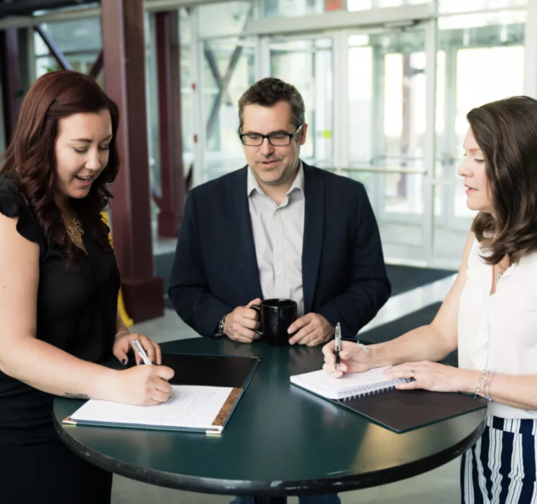 Three professionals with documents at a round table in a modern office setting.