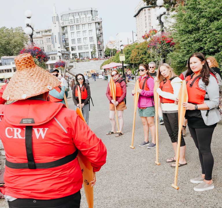 Group of people wearing life jackets and holding paddles on a city street with crew member in foreground.